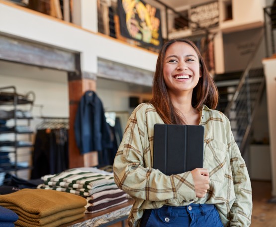 a woman stands in a small business with a warm smile while holding a tablet 