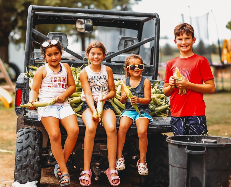 a group of kids sit on a 4x4 shucking corn