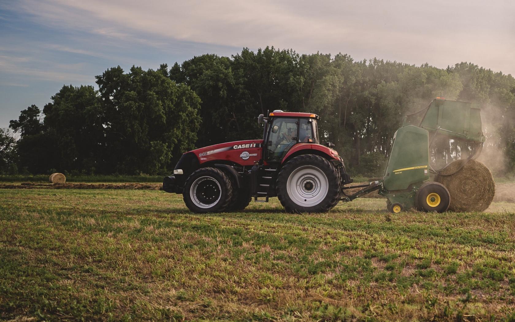 A red tractor in a field baling hay with a green round baler